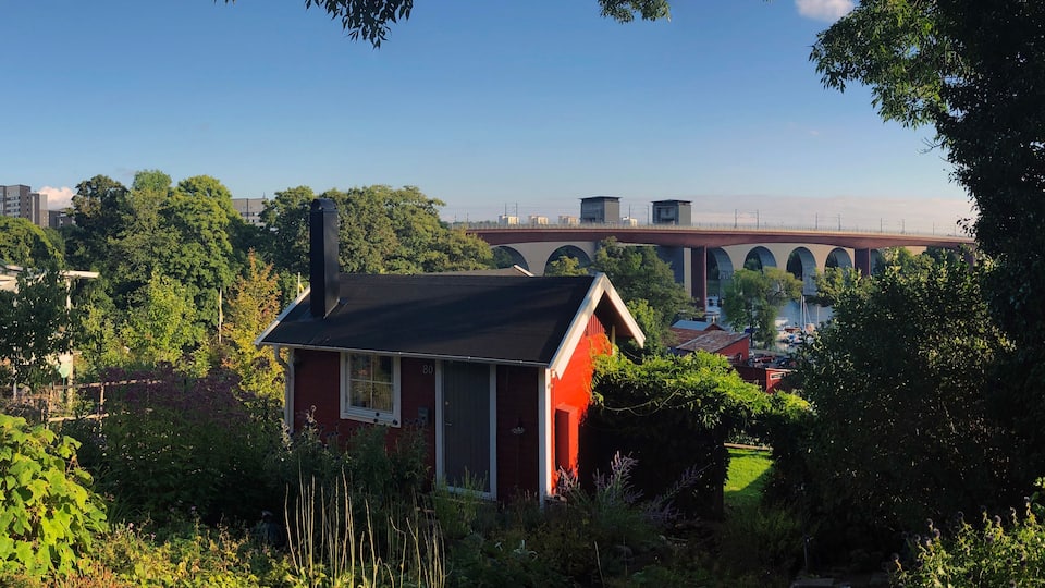 A red house with a black roof surrounded by lush greenery, a bridge, and residential buildings in the background.