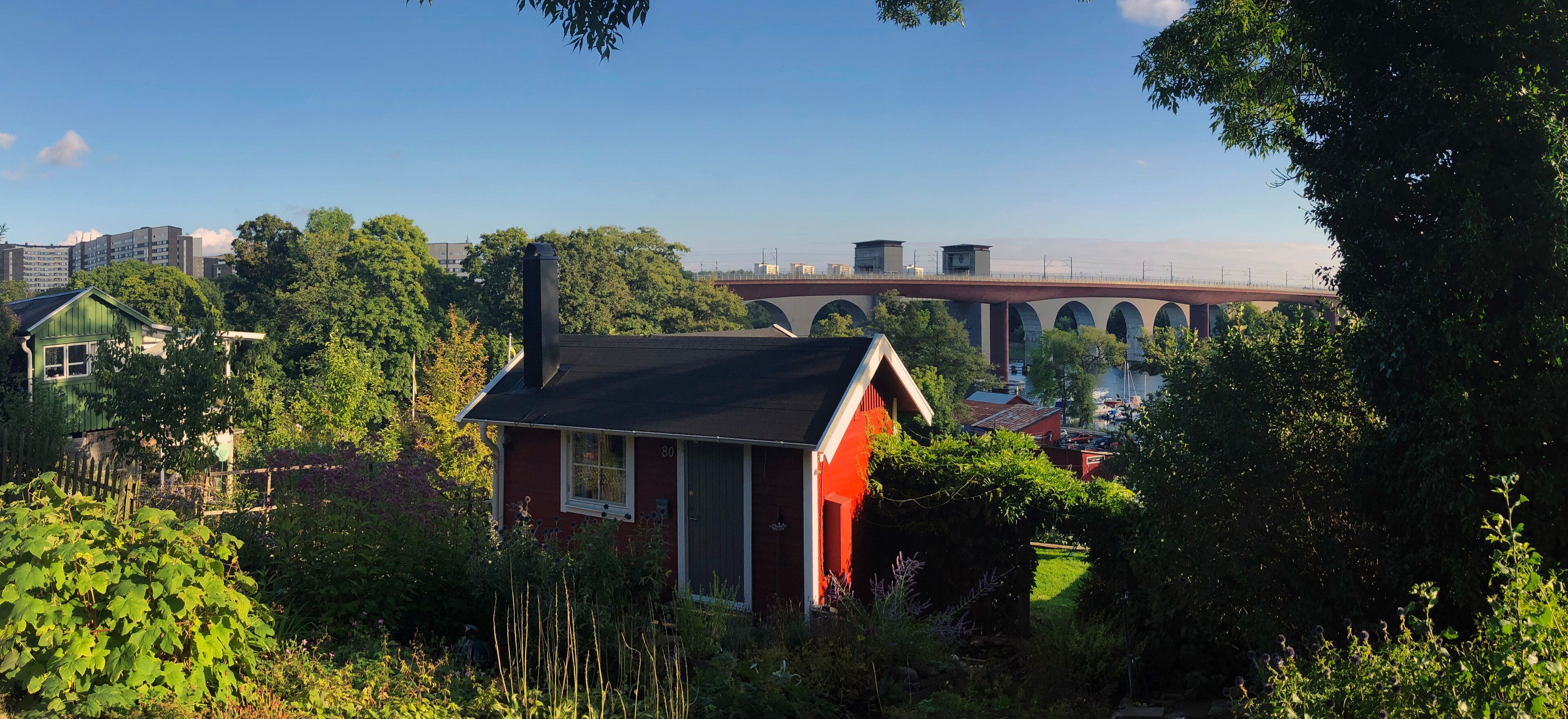 A red house with a black roof surrounded by lush greenery, a bridge, and residential buildings in the background.