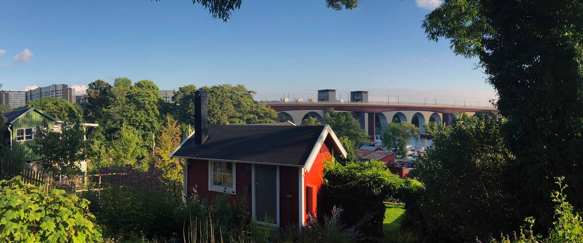 A red house with a black roof surrounded by lush greenery, a bridge, and residential buildings in the background.