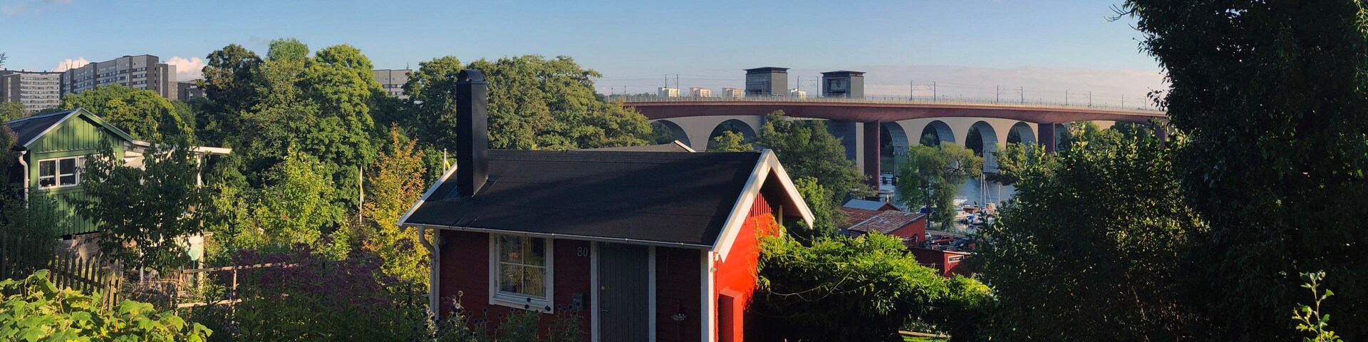 A red house with a black roof surrounded by lush greenery, a bridge, and residential buildings in the background.