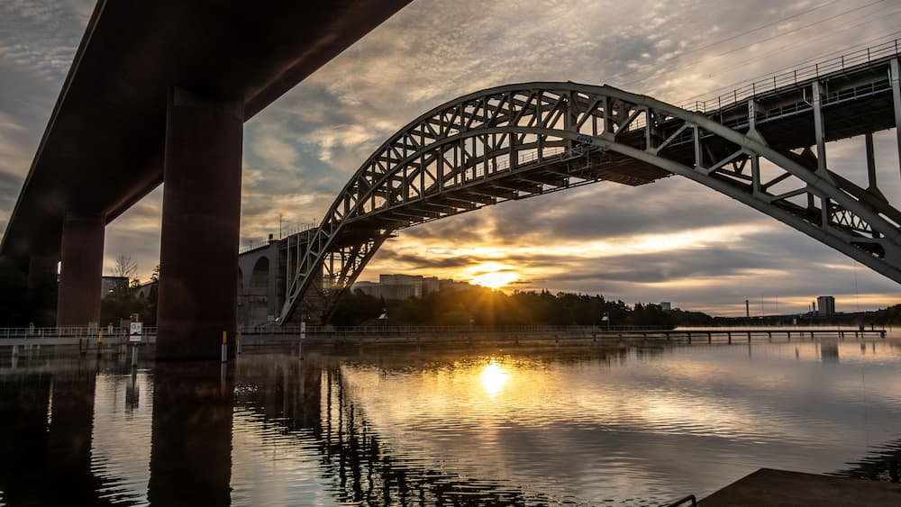 Un pont en acier, dont l’arche s’étend au-dessus d’une rivière paisible, avec le soleil couchant en arrière-plan.