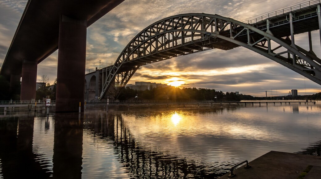 A steel bridge arching over a calm river with the sun setting in the background.