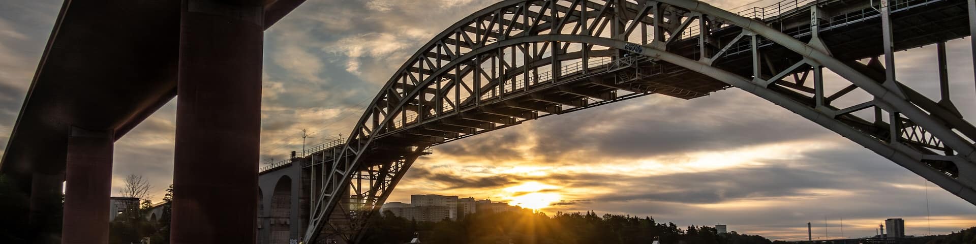 A steel bridge arching over a calm river with the sun setting in the background.