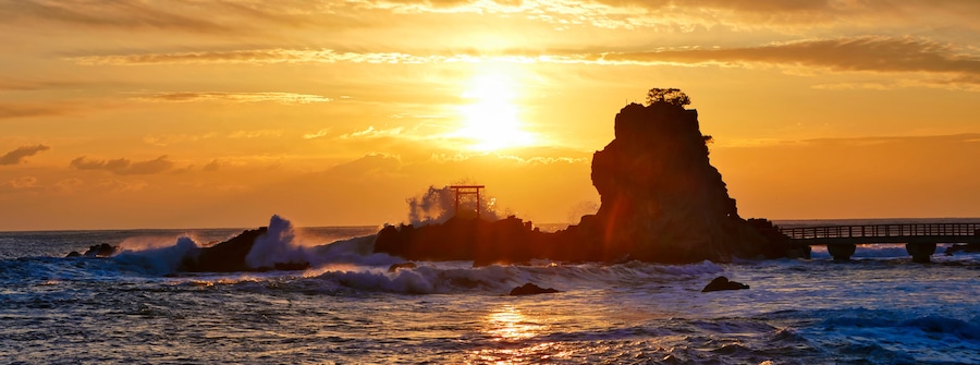 A sunset over a rocky coastline with waves crashing against the rocks and a bridge in the background.