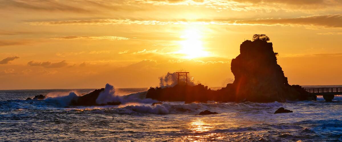 A sunset over a rocky coastline with waves crashing against the rocks and a bridge in the background.