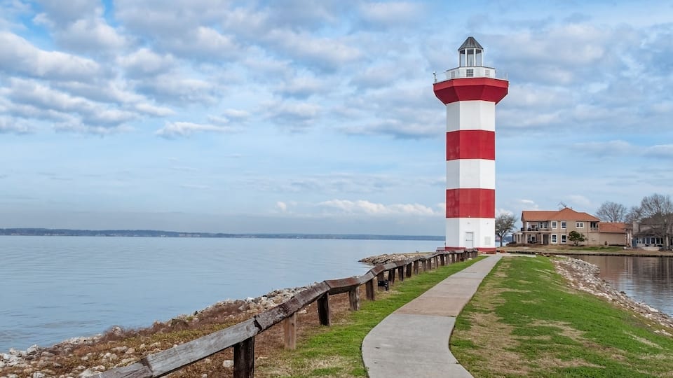 A red and white lighthouse by a calm body of water with a wooden walkway and a house in the background.