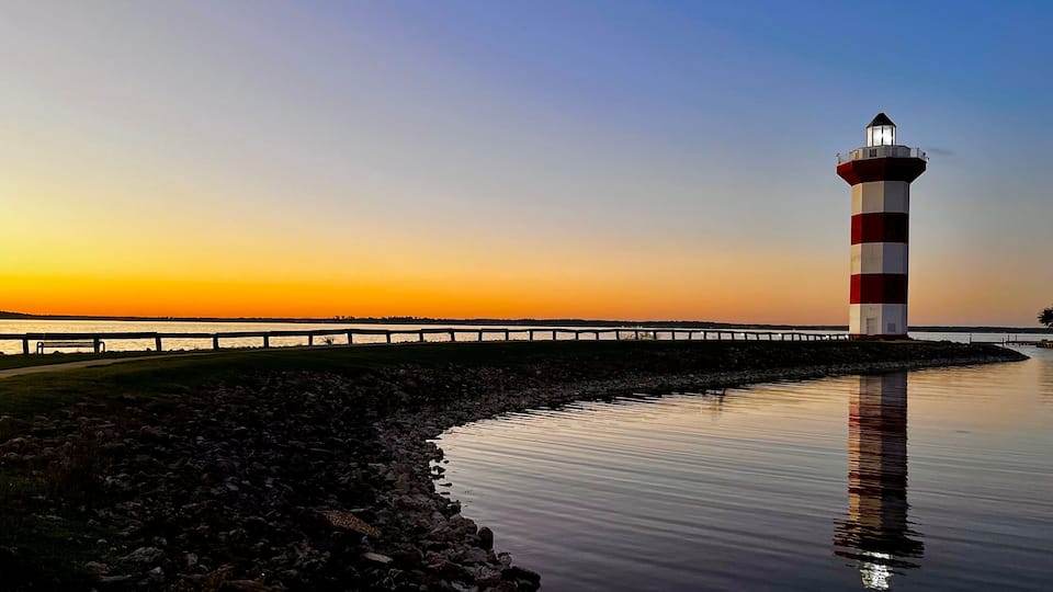 A lighthouse with red and white stripes, a calm body of water reflecting the structure, a wooden walkway, and a sunset sky.