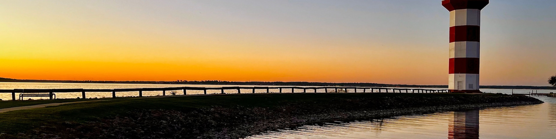 Un faro con franjas rojas y blancas, una masa de agua tranquila que refleja la estructura, un pasillo de madera y un cielo al atardecer.