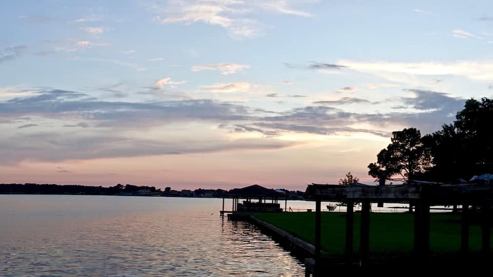 A calm lake at sunset with a pier on the right and trees on the left.