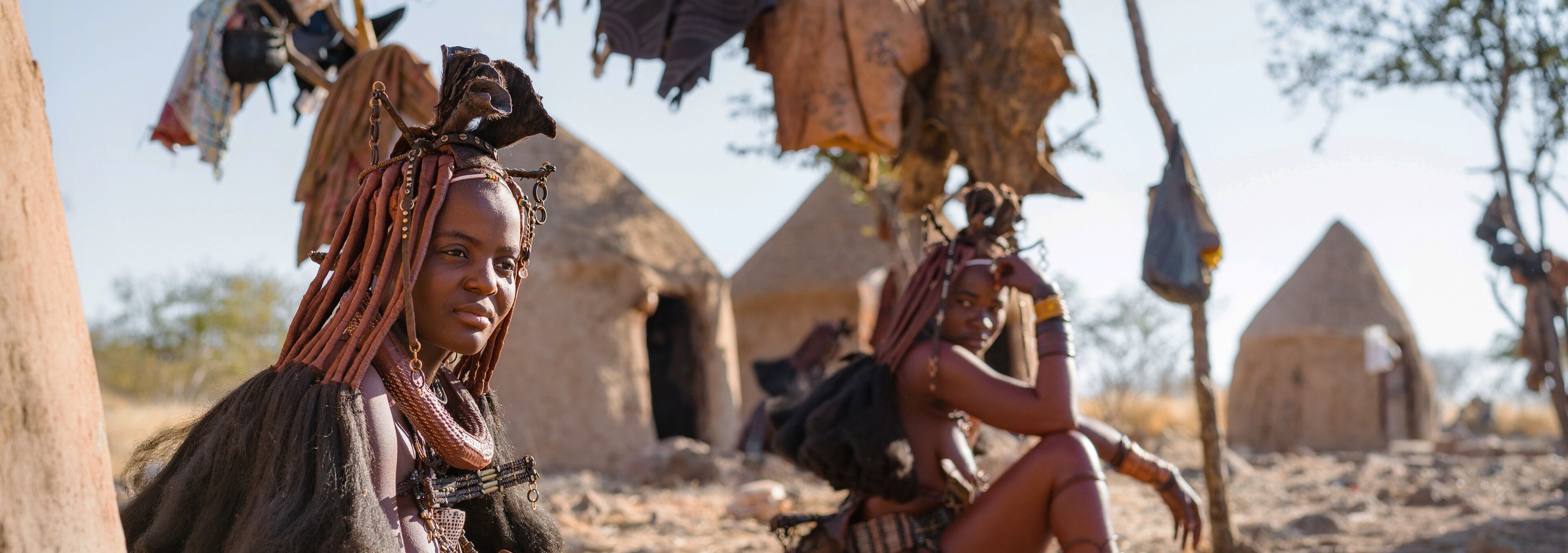 A person with braided hair and a necklace sitting in front of a traditional hut.