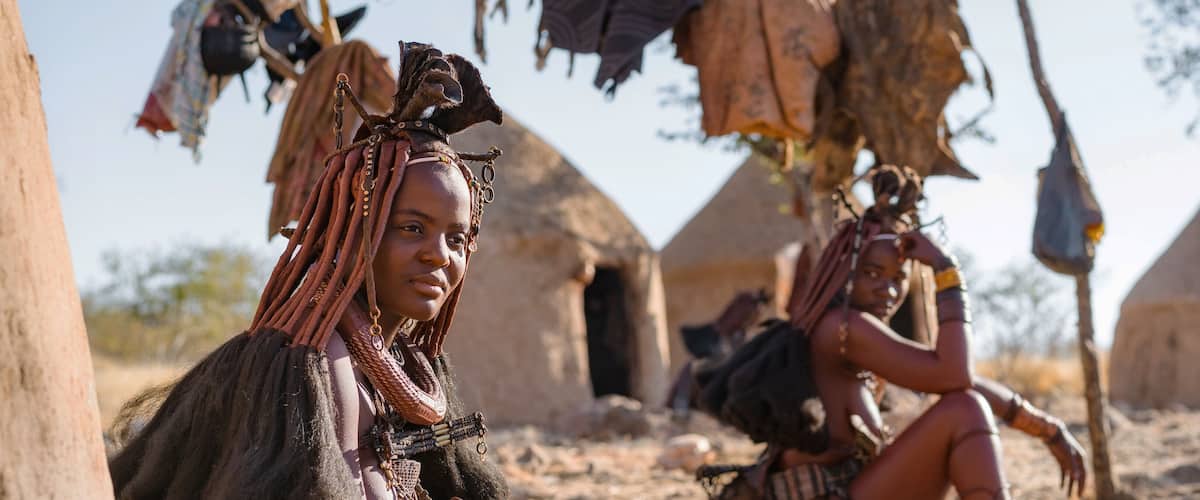 A person with braided hair and a necklace sitting in front of a traditional hut.