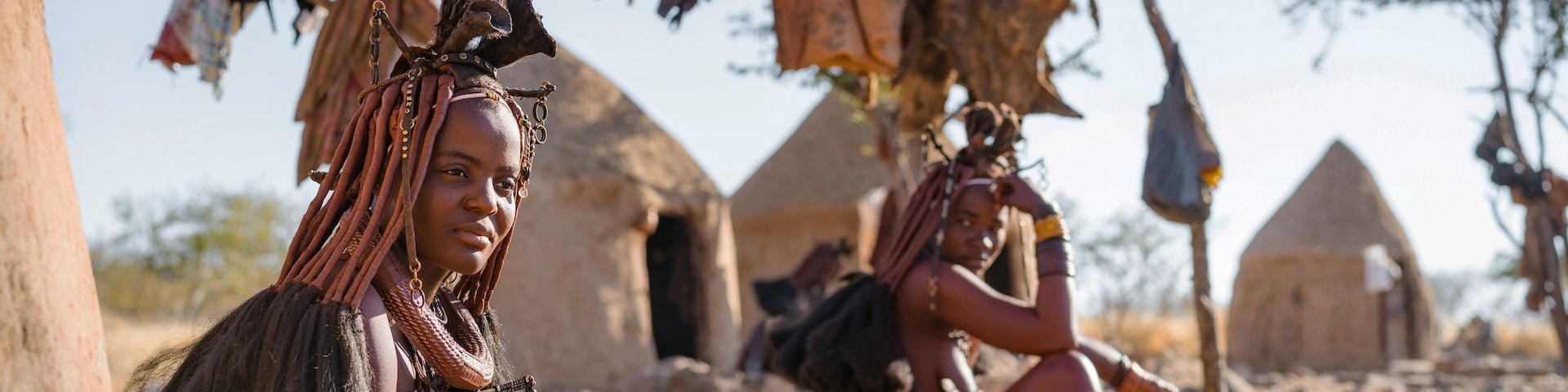 A person with braided hair and a necklace sitting in front of a traditional hut.