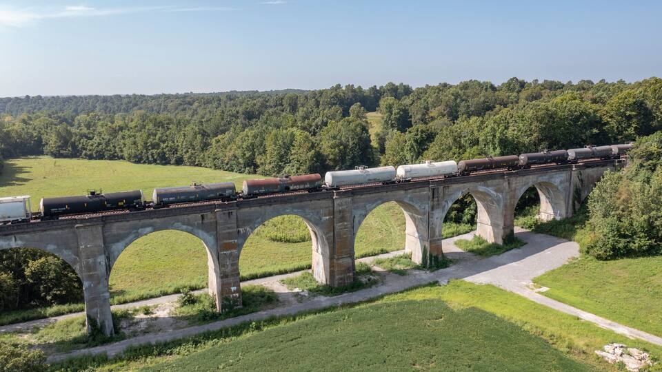 A train crossing a stone bridge over a river, surrounded by lush greenery.