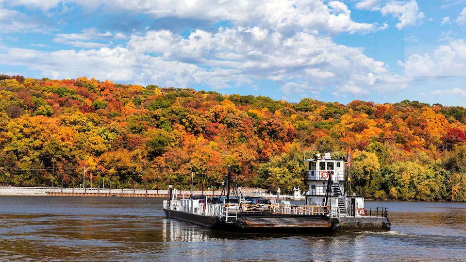 A ferry boat on a river with a backdrop of colorful autumn foliage.