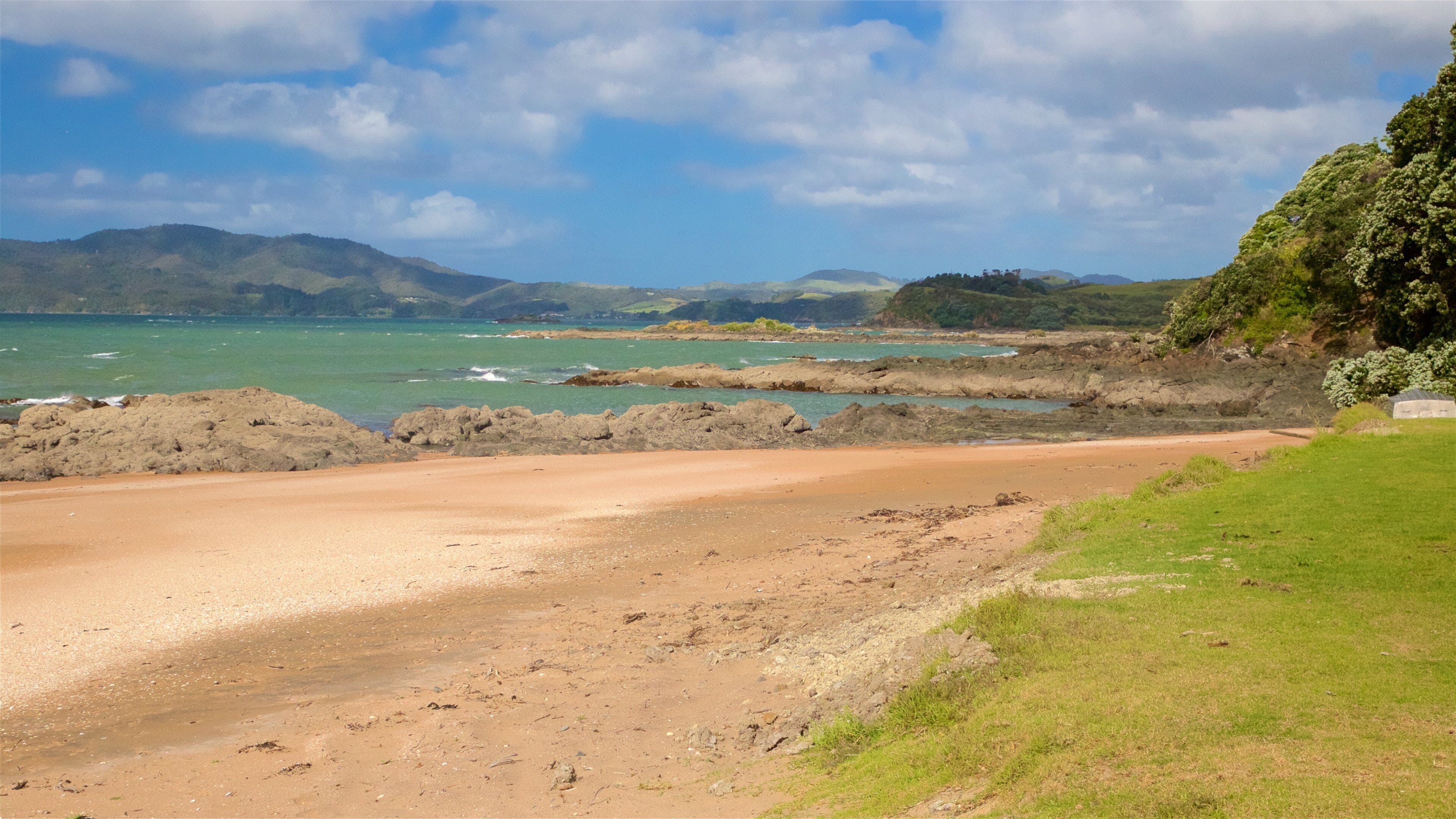 A coastal landscape with a sandy beach, rocky outcrops, and distant mountains under a partly cloudy sky.