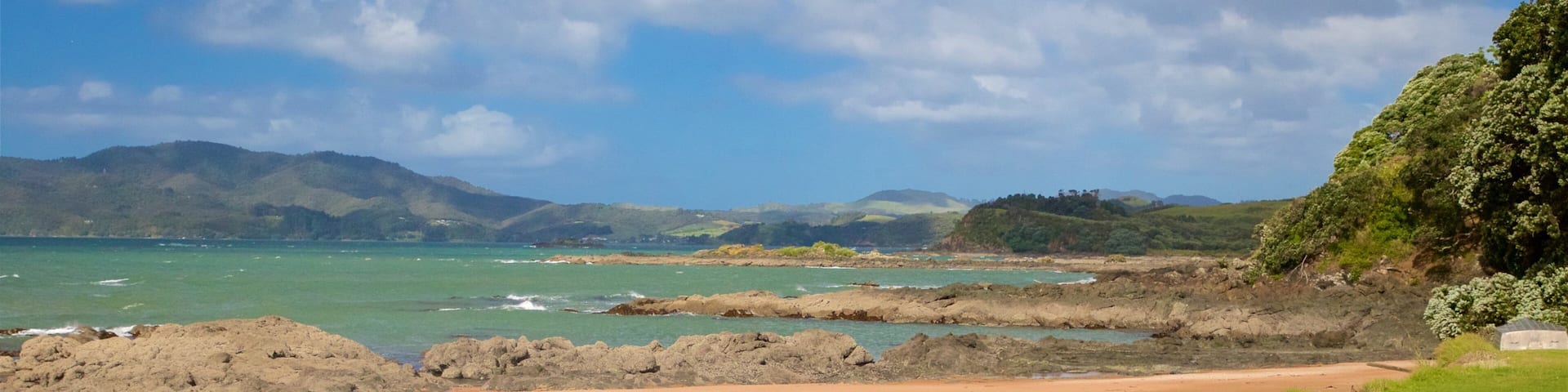 A coastal landscape with a sandy beach, rocky outcrops, and distant mountains under a partly cloudy sky.