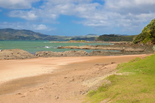 A coastal landscape with a sandy beach, rocky outcrops, and distant mountains under a partly cloudy sky.