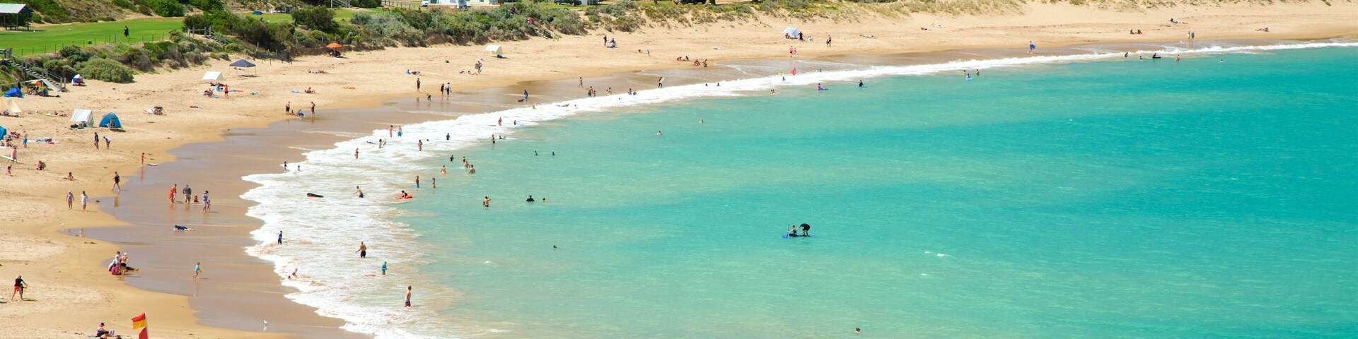 A crowded beach with clear turquoise water, sandy shores, and a backdrop of greenery and buildings.