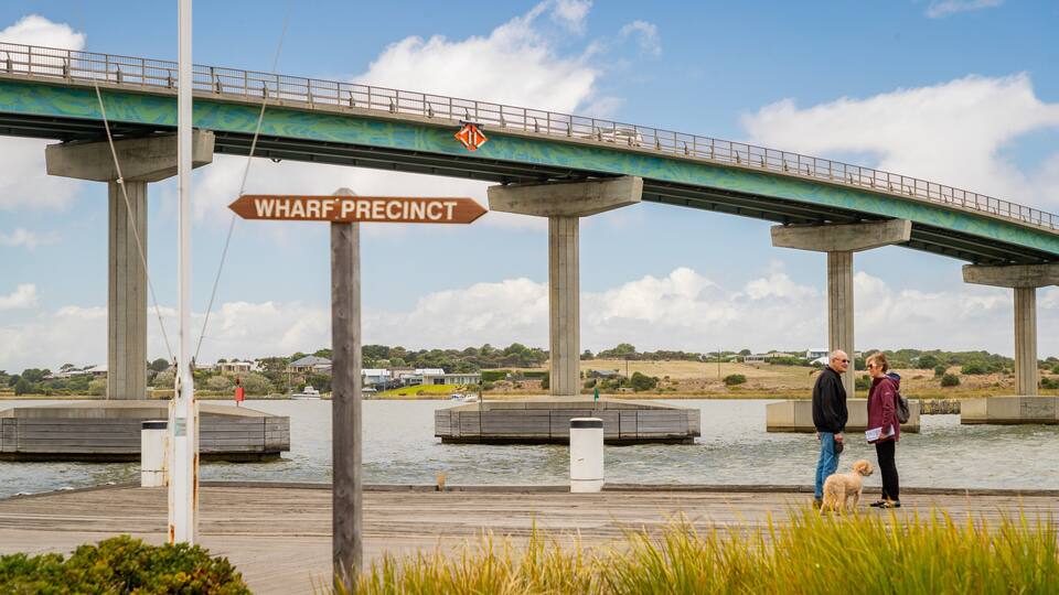 A bridge with a sign reading "WHARF PRECINCT" and a wooden dock with people and a dog nearby.
