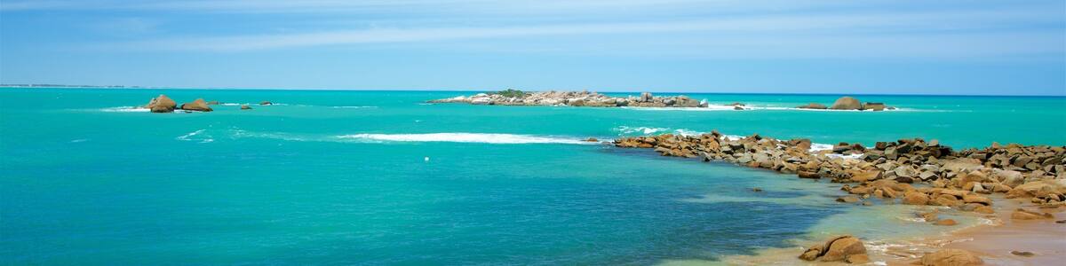 Une plage bordée d’une eau turquoise et limpide, de formations rocheuses et d’une île visible au loin.
