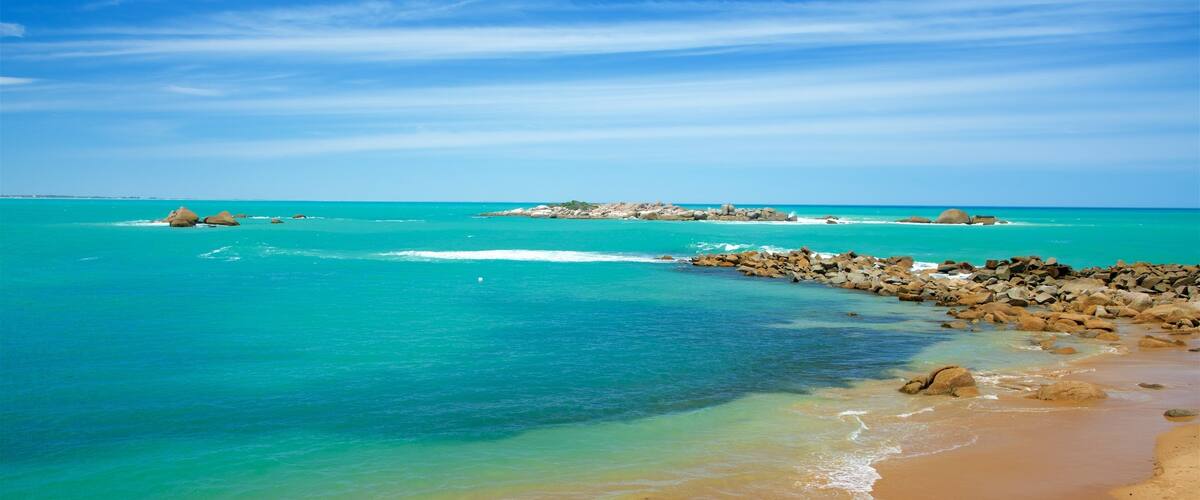 Una playa con agua turquesa cristalina, afloramientos rocosos y una isla distante.