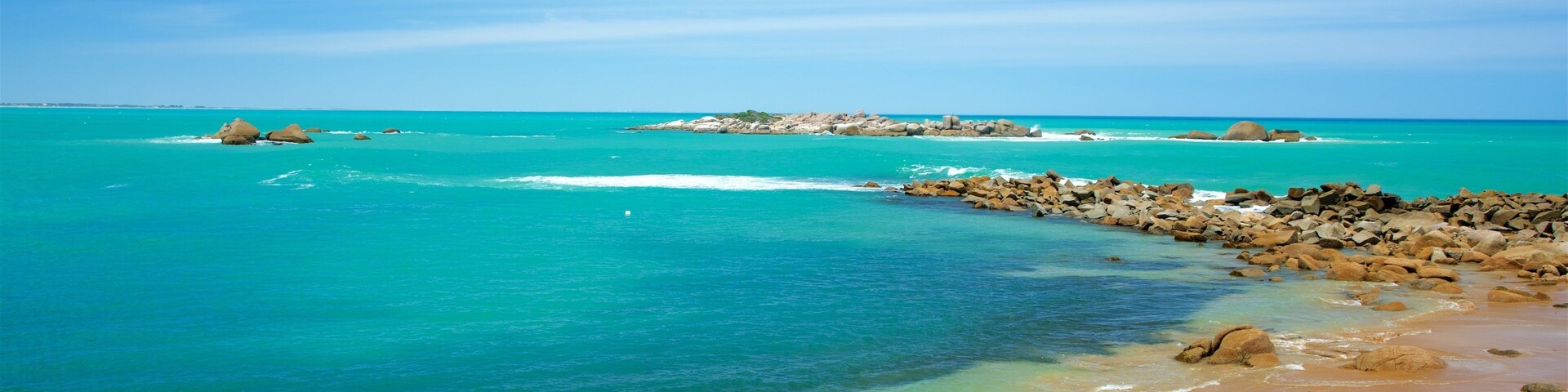 Una playa con agua turquesa cristalina, afloramientos rocosos y una isla distante.