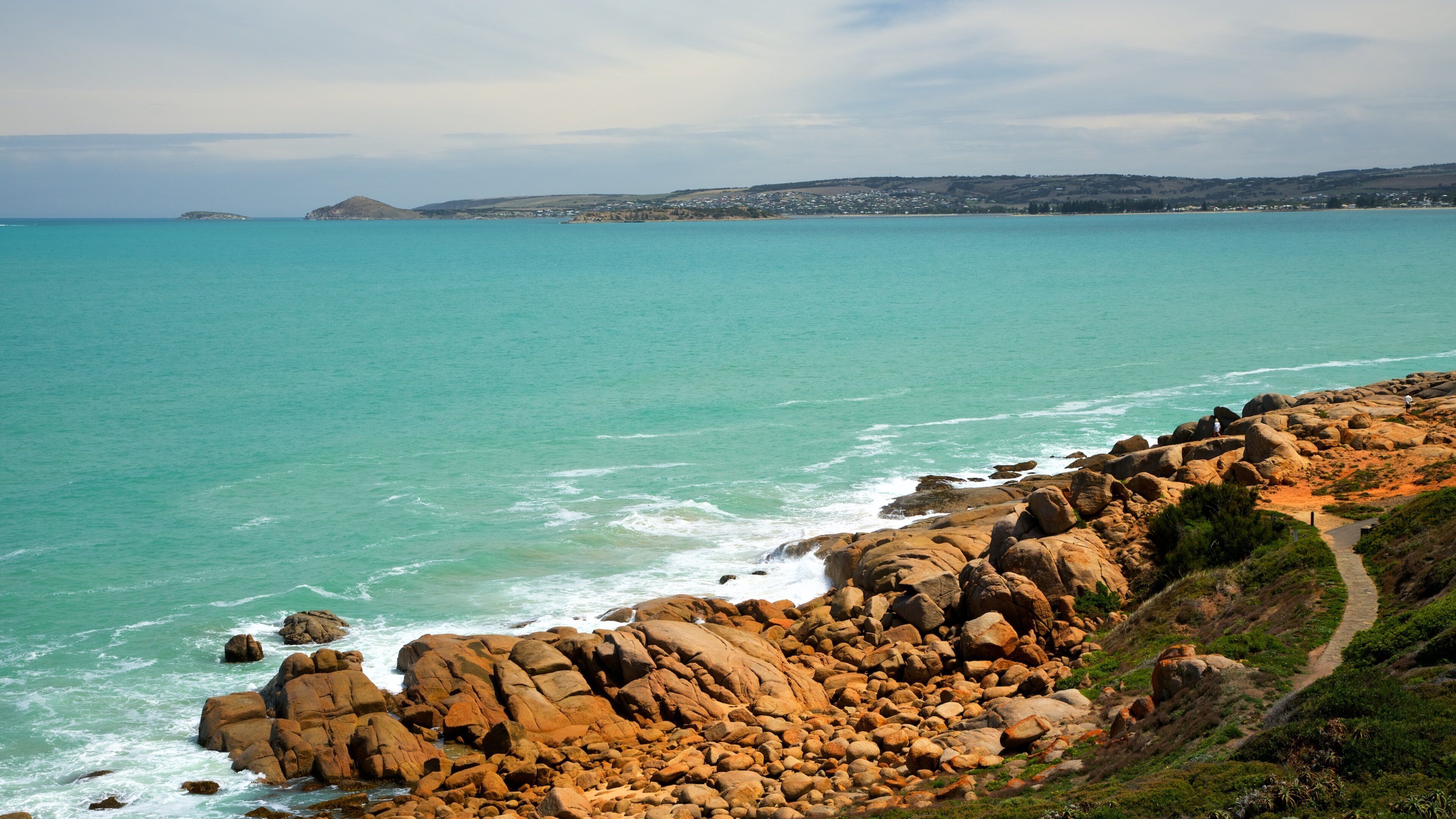 A coastal landscape with rocky cliffs, turquoise sea, and a distant town.