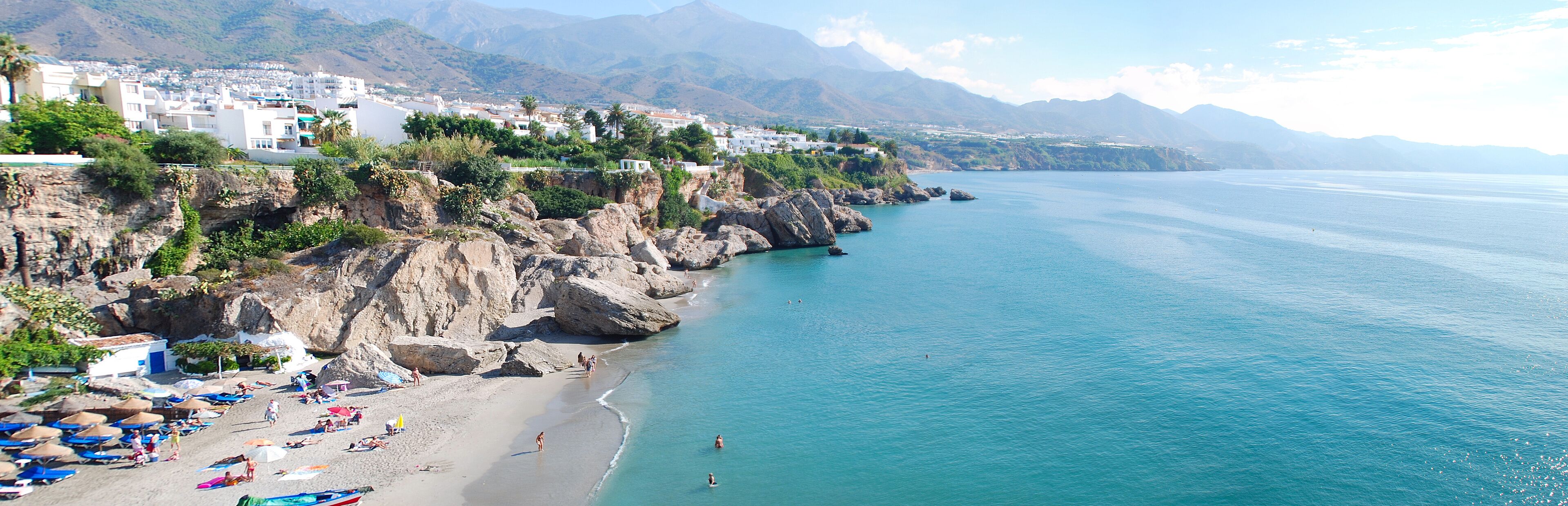 A coastal town with a beach, rocky cliffs, and mountains in the background.