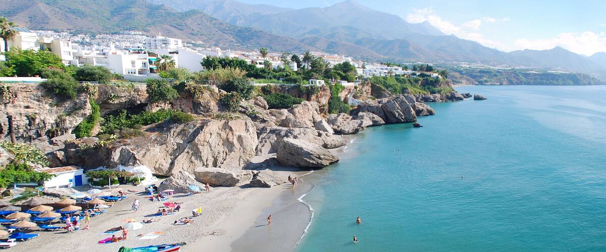 A coastal town with a beach, rocky cliffs, and mountains in the background.