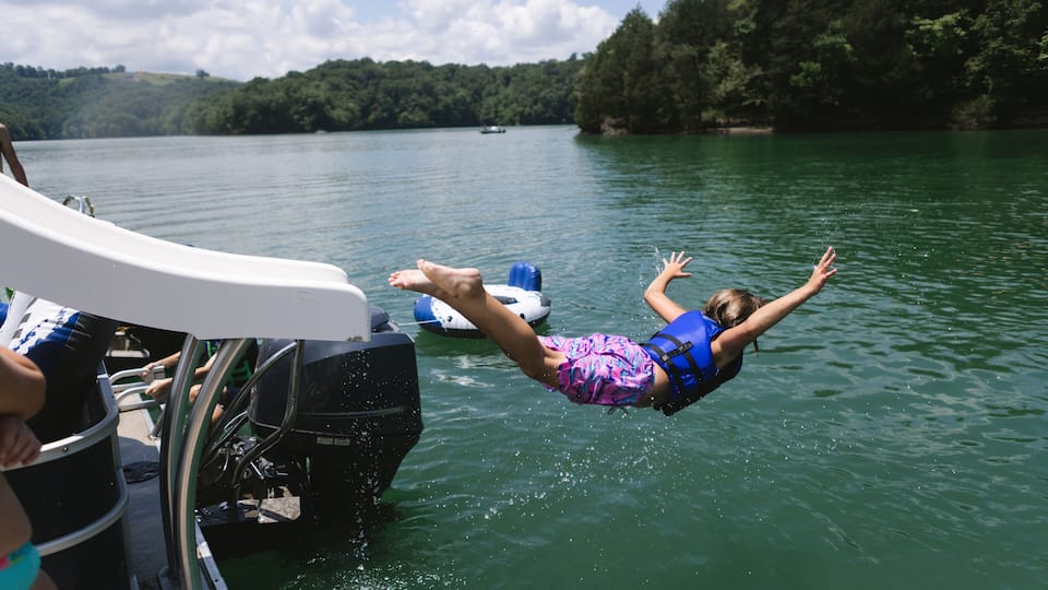 A person mid-air off a boat, water splashing around, with a forested shoreline and a boat in the background.