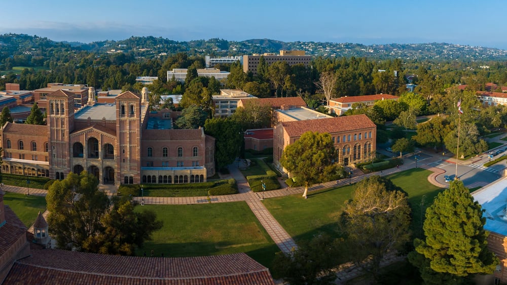 Aerial view of UCLA campus with Gothic tower, red-brick buildings, green lawns, and pathways amidst hilly, tree-covered landscape in soft, golden light.