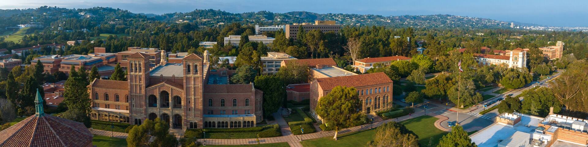 Aerial view of UCLA campus with Gothic tower, red-brick buildings, green lawns, and pathways amidst hilly, tree-covered landscape in soft, golden light.