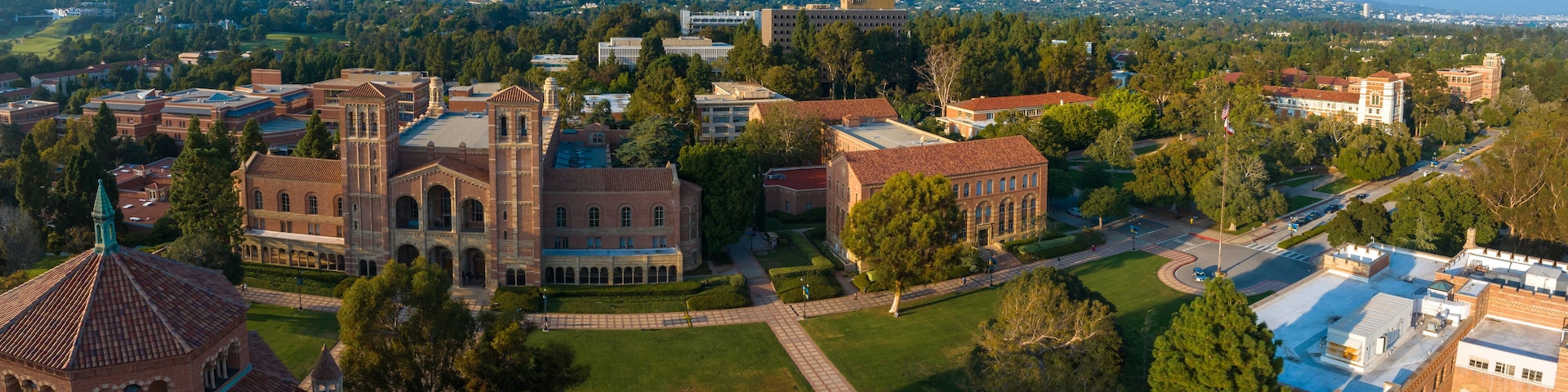 Aerial view of UCLA campus with Gothic tower, red-brick buildings, green lawns, and pathways amidst hilly, tree-covered landscape in soft, golden light.