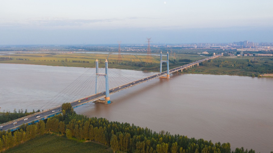 A large suspension bridge spanning a wide river, surrounded by green fields and a distant city skyline.