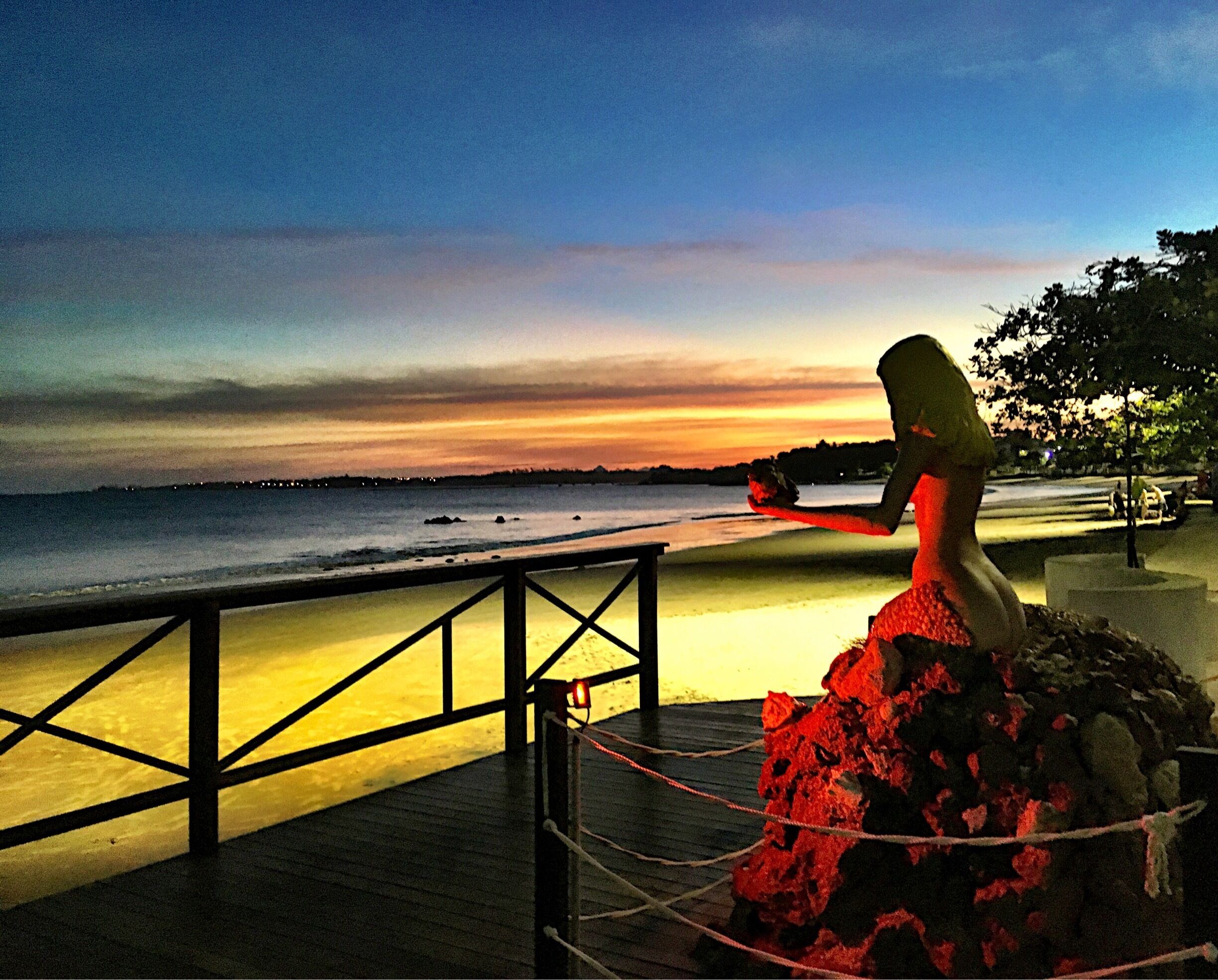 A sunset over a beach with a statue of a woman holding a shell.