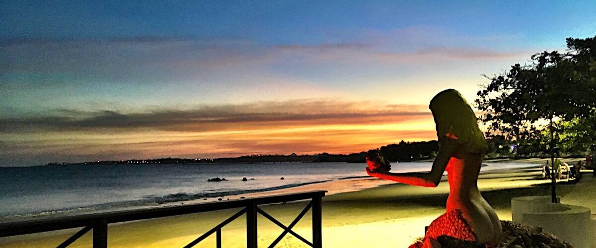 A sunset over a beach with a statue of a woman holding a shell.