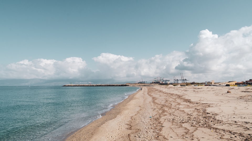 A beach with a rocky breakwater, calm sea, and industrial structures in the distance.