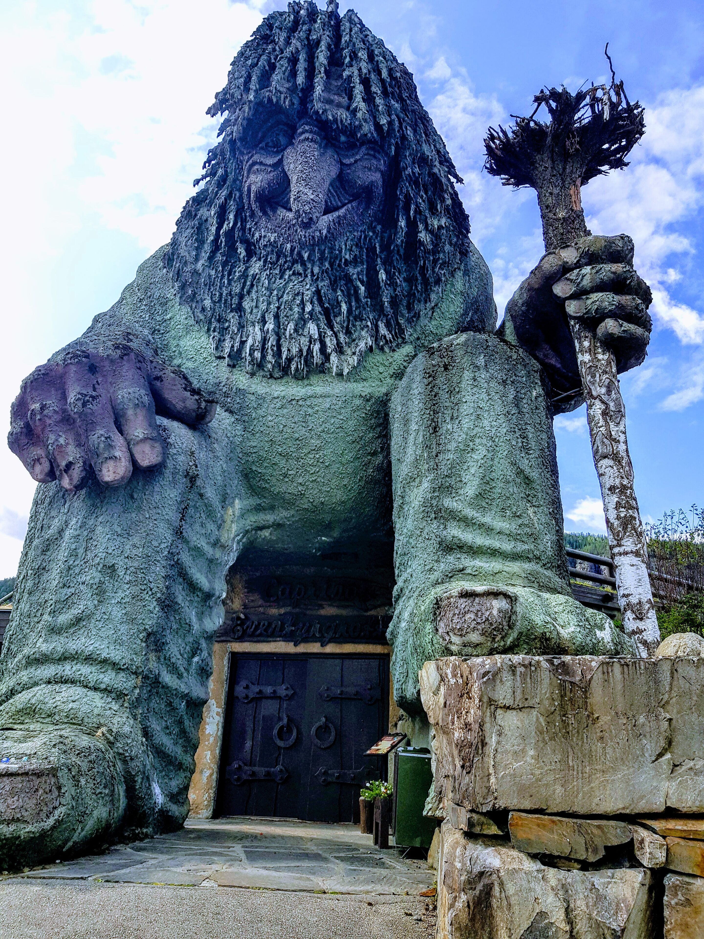 A large stone statue of a figure with a beard and a staff, sitting in front of a black door.