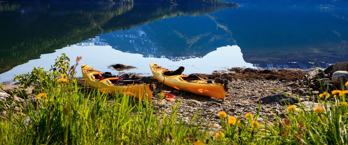 A calm lake with kayaks on the shore, surrounded by mountains and greenery.
