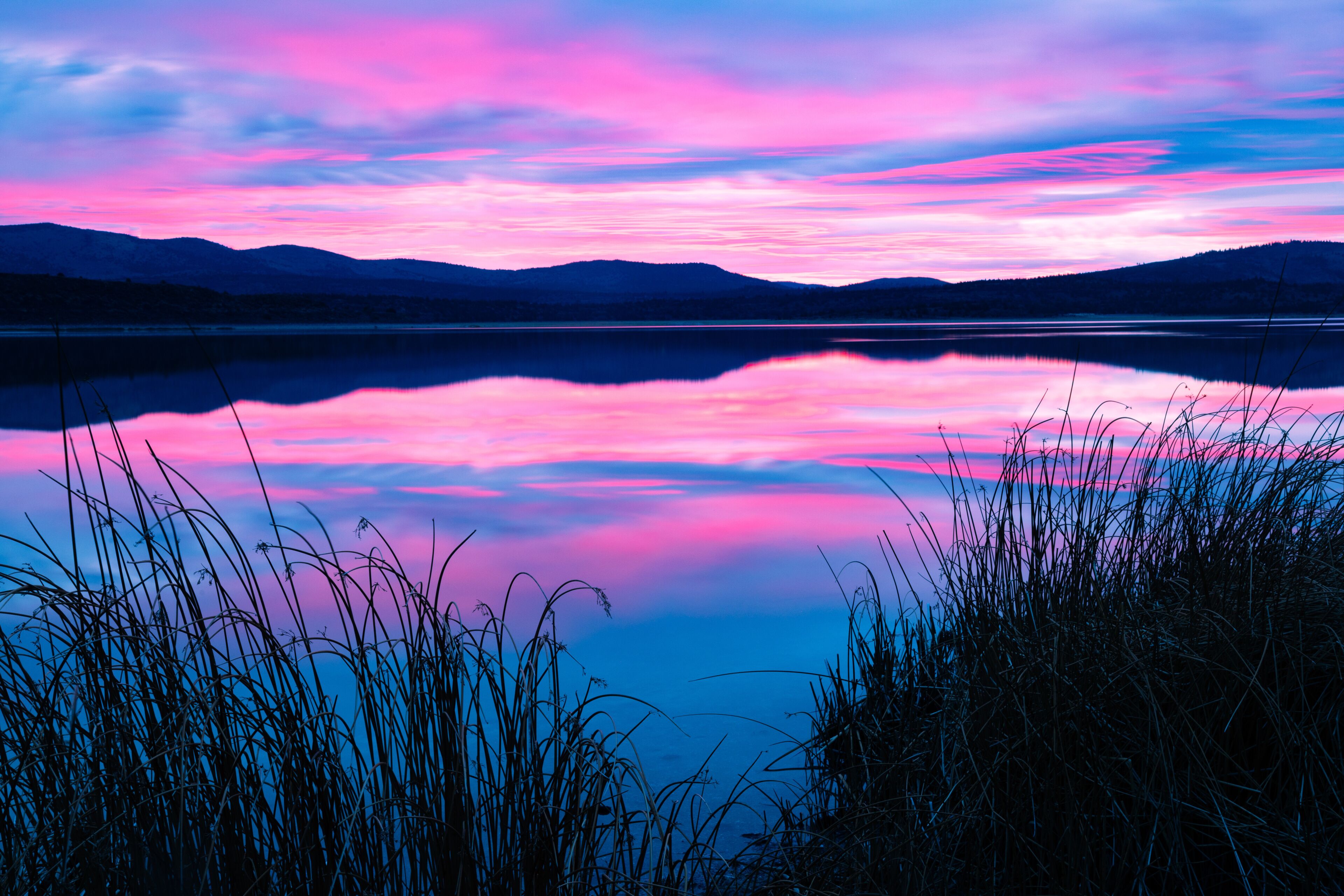 A calm lake with a reflection of the sky, surrounded by grass and mountains.