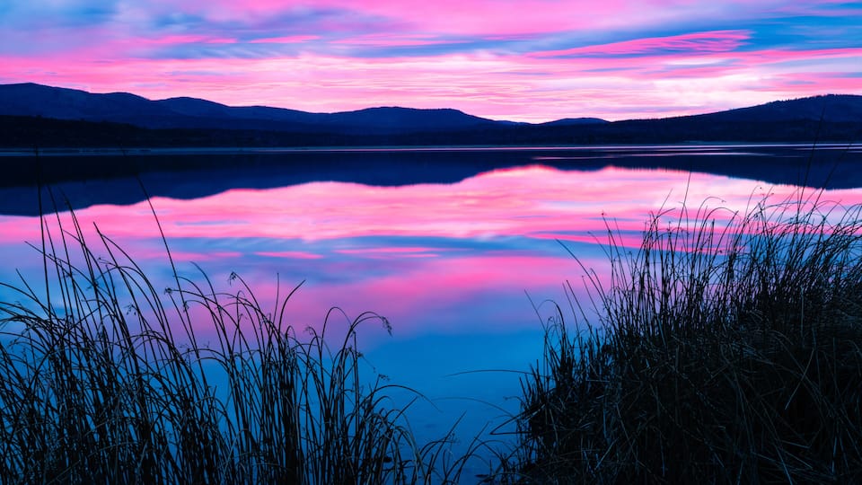A calm lake with a reflection of the sky, surrounded by grass and mountains.