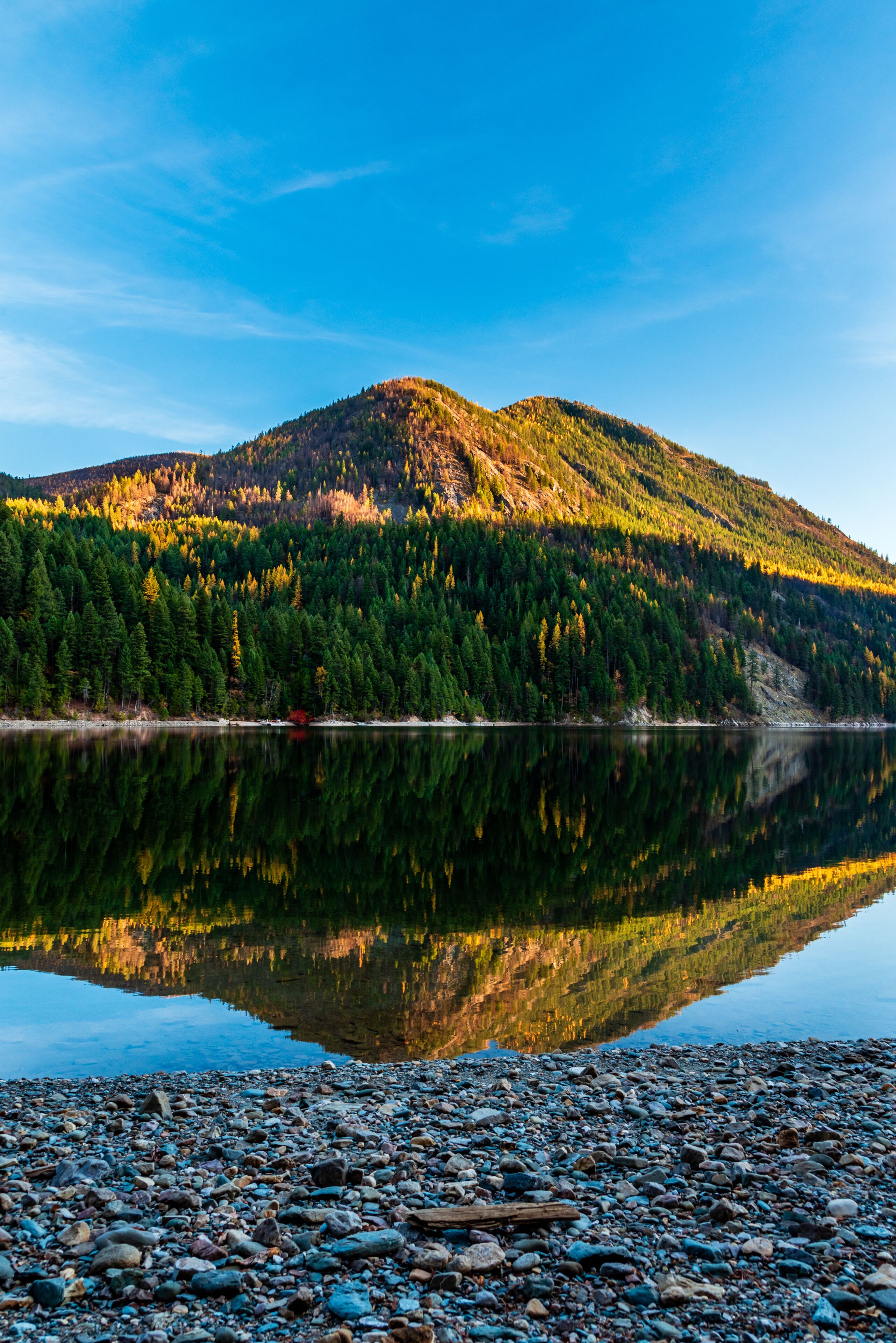 A calm lake reflecting a forested mountain, a rocky shoreline, and a clear blue sky.