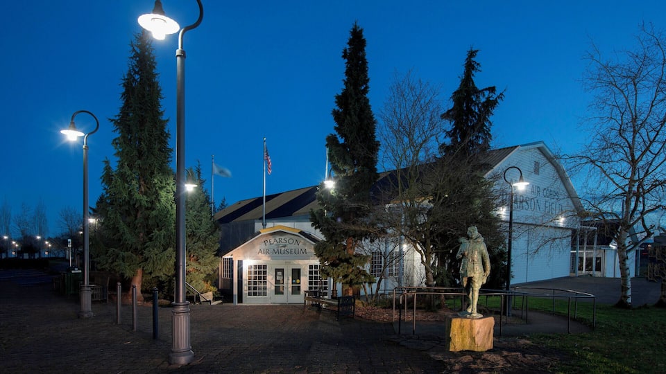 The Pearson Air Museum at dusk with illuminated street lamps and a statue in the foreground.