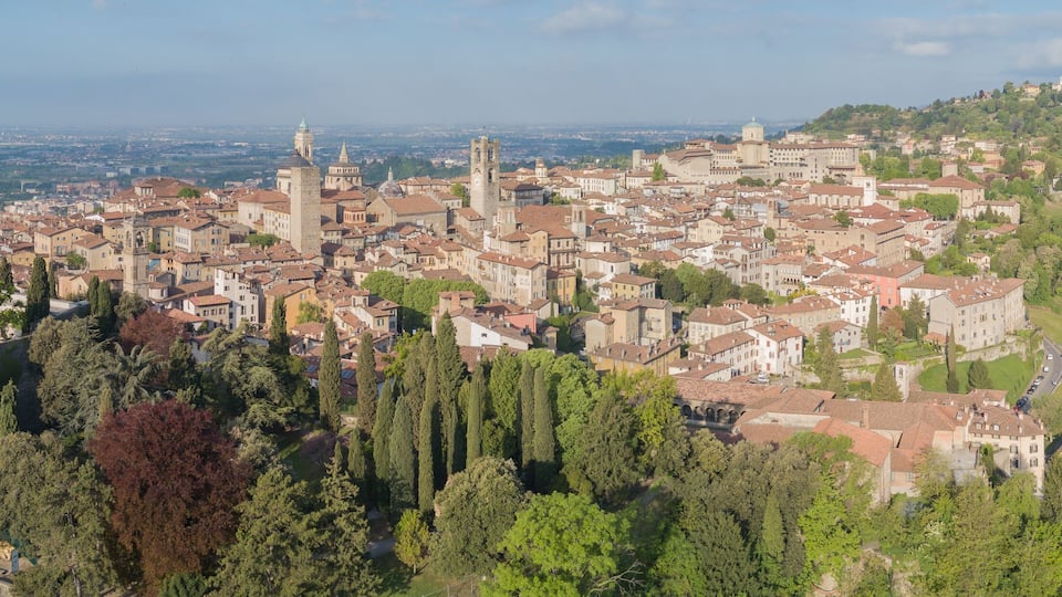 A panoramic view of a historic town with medieval architecture, including towers and a prominent church, surrounded by lush greenery and hills.