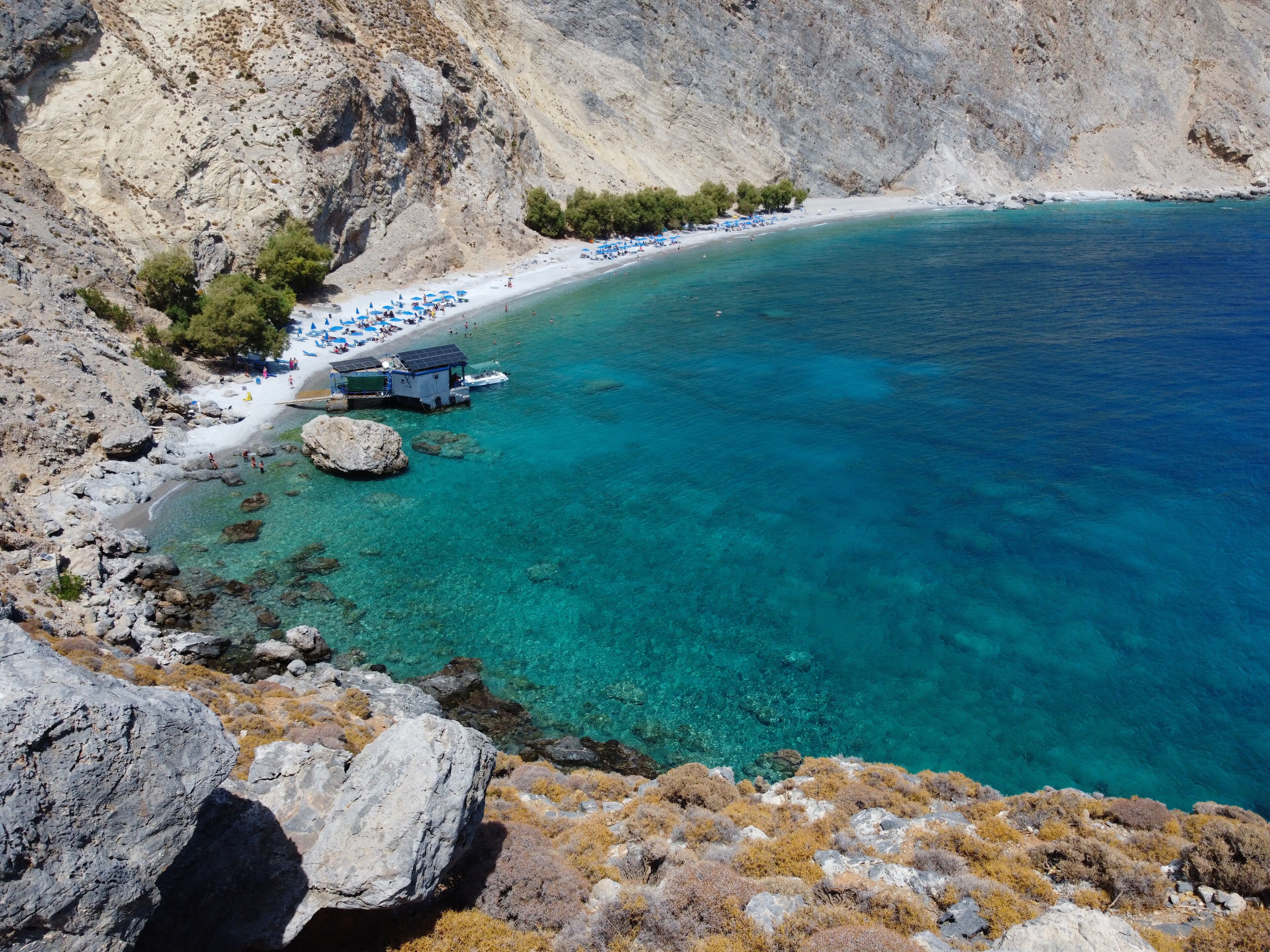 A coastal landscape with clear turquoise waters, rocky terrain, and a beach with umbrellas.