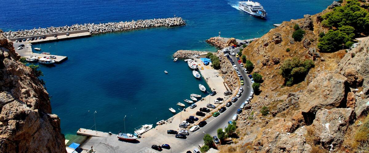 A coastal road with parked cars, a pier, boats, and a cruise ship.