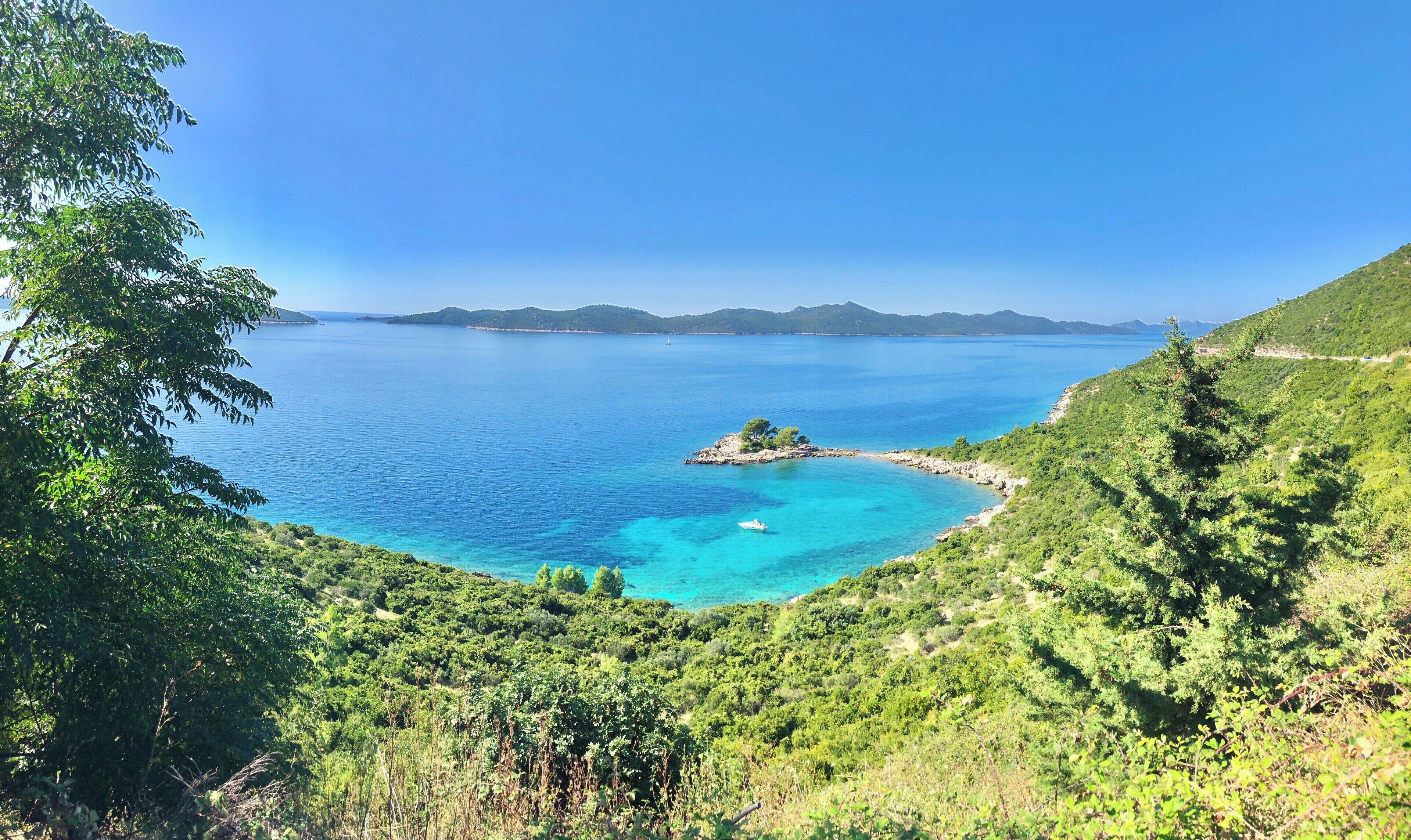 A coastal landscape with a small island surrounded by clear blue waters, dense green vegetation, and distant mountains.