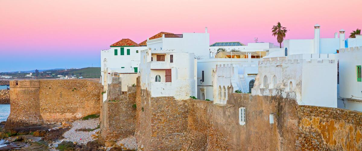 A coastal town with white buildings, a stone wall, and a pink sky.