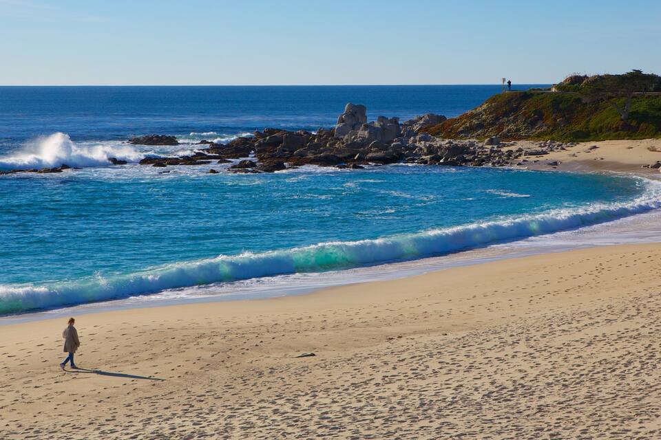 Uma praia arenosa com ondas quebrando na costa, um afloramento rochoso e uma pessoa caminhando.