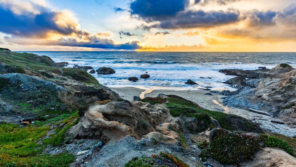 A rocky coastline with waves crashing against the shore under a dramatic sunset sky.